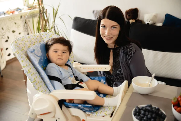 Madre alimentando a su hijo gachas en el asiento del niño - Niño asiático de etnia mixta con camisa de cuerpo azul con dibujos animados oso sonriente Embarazada — Foto de Stock
