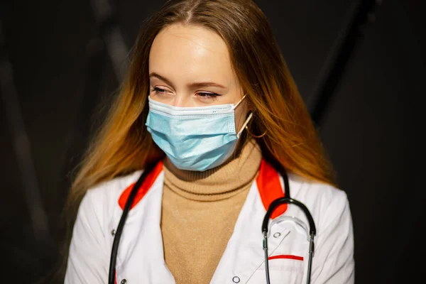 Retrato de médico ou enfermeira feliz em máscara cirúrgica e vestido médico branco olhando para você com o vento pegando cabelo — Fotografia de Stock