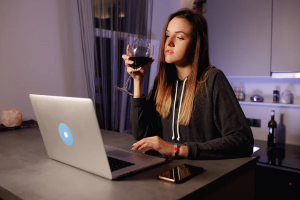 Genç bir kadın akşam karantina sırasında dizüstü bilgisayarda oturur ve kırmızı şarap içer.