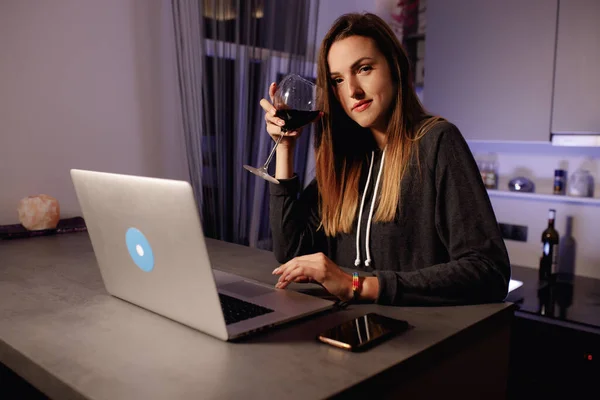 Güzel bir kadın laptopta çalışıyor ve akşam karantina sırasında kameraya bakarak kırmızı şarap içiyor.