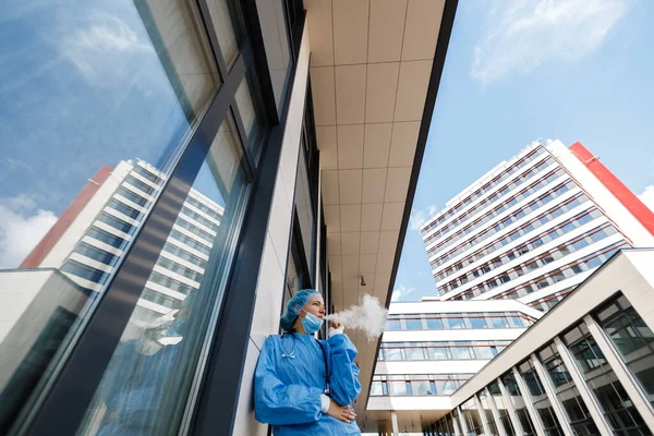 Wide low angle: Young doctor or nurse on break smokes electronic cigarette wearing surgical face mask, cap and gown near hospital