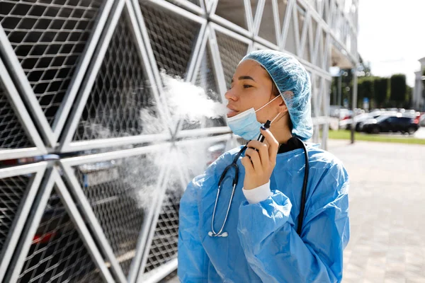 Fáradt fiatal orvos vagy nővér szünetben műtét után dohányzik elektronikus cigaretta visel sebészeti arc maszk, sapka és köpeny közelében kórház — Stock Fotó