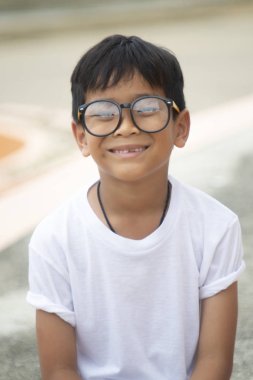 Asyalı çocuk gözlüklü beyaz bir t-shirt giyiyor, oturan, mutlu gülümseyen