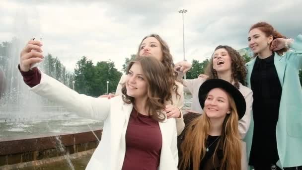 Selfies al aire libre en la ciudad. Cinco chicas toman fotos en un smartphone junto a la fuente. jóvenes. concepto de turismo. Feliz verano divertido — Vídeo de stock