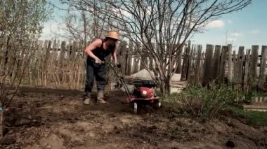 Genç ve güçlü bir adam bahçedeki ekiciye sürtünüyor. Tohum ekimi için toprak hazırlığı. Çiftlikte çalış. Tarım ve sıkı çalışma kavramı. Traktör sürücüsü. Saban. 30-40 yaşlarında.