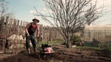 Genç ve güçlü bir adam bahçedeki ekiciye sürtünüyor. Tohum ekimi için toprak hazırlığı. Çiftlikte çalış. Tarım ve sıkı çalışma kavramı. Traktör sürücüsü. Saban. 30-40 yaşlarında.