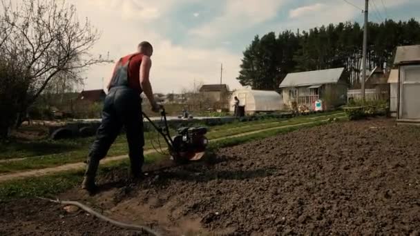 Genç ve güçlü bir adam bahçedeki ekiciye sürtünüyor. Tohum ekimi için toprak hazırlığı. Çiftlikte çalış. Tarım ve sıkı çalışma kavramı. Traktör sürücüsü. Saban. 30-40 yaşlarında. — Stok video