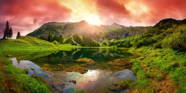 山のあるアルプスの湖での劇的な日の出 澄んだ水と鮮やかな緑の草を導くパスに反映された赤い雲 — ストック写真