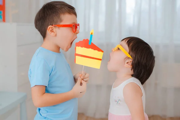 Menino e uma menina brincando com adereços bolo de papel com uma vela. Desempenho das crianças de morder o bolo juntos em seu aniversário . — Fotografia de Stock