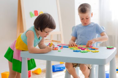 Bir erkek ve bir kız masada yumuşak bir yapboz topluyor. Erkek ve kız kardeş odada birlikte oynarken eğleniyorlar. Anaokulu çocukları ve eğitici oyuncaklar