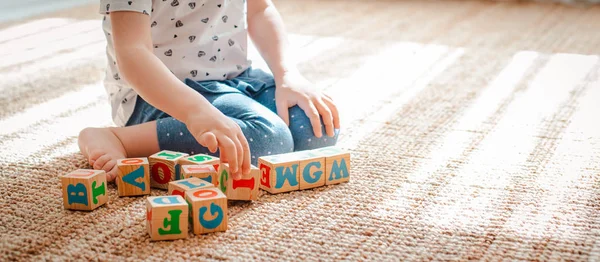 Kind spielt mit Holzklötzen mit Buchstaben auf dem Boden im Zimmer ein kleines Mädchen baut zu Hause oder im Kindergarten einen Turm. — Stockfoto