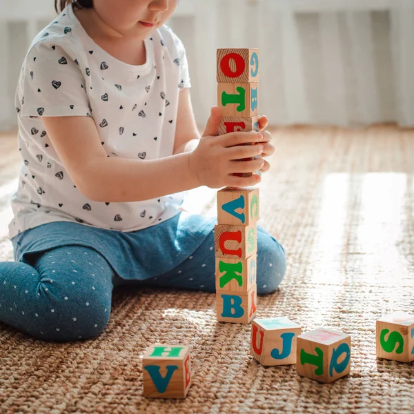 Niño juega con bloques de madera con letras en el suelo en la habitación una niña está construyendo una torre en casa o en el jardín de infantes . — Foto de Stock