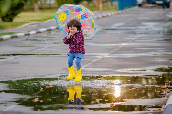 Happy funny child with multicolored umbrella jumping puddles in rubber boots and laughing — Stock Photo, Image