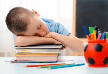 Okullu çocuk sınıfında oturmuş kitap dolu bir masada yatıyor. Uyuyan tembel öğrencileri eğitiyor.