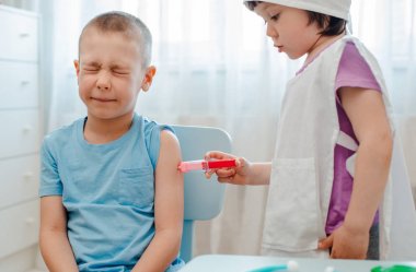 Çocuklar hastanede oynuyor. Kız doktor gibi giyinmiş. 4 yaşında bir çocuğa enjeksiyon yapıyor. 6 yaşında bir çocuğa. Anaokulundaki bir odada oyuncak şırıngayla..