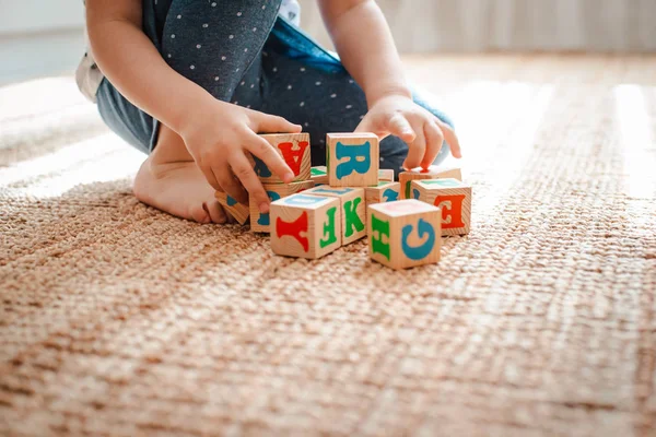 Child plays with wooden blocks with letters on the floor in the room a little girl is building a tower at home or in the kindergarten. — Stock Photo, Image