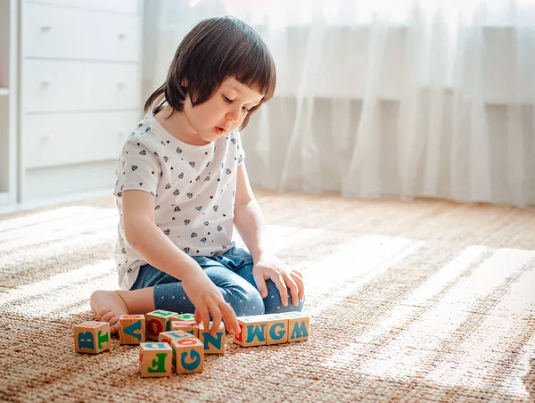 Enfant joue avec des blocs de bois avec des lettres sur le sol dans la chambre une petite fille construit une tour à la maison ou dans la maternelle . — Photo