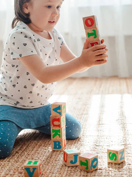 Barn leker med träklossar med bokstäver på golvet i rummet en liten flicka bygger ett torn hemma eller i dagis. — Stockfoto