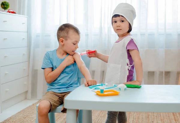 Τα παιδιά παίζουν στο νοσοκομείο. Το κορίτσι είναι ντυμένο σε ένα γιατρό 4 ετών κάνει μια ένεση σε ένα αγόρι 6 ετών με μια σύριγγα παιχνιδιού σε ένα δωμάτιο σε ένα νηπιαγωγείο. — Φωτογραφία Αρχείου