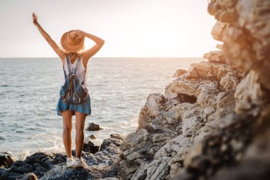 Şapkalı genç bir hipster kadın ve elleri havada bir rukzak, bir uçurumun tepesinde duran ve gün batımında denize bakıyor.