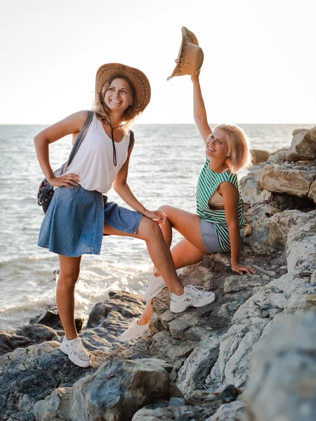 หญิงสาวสองคนที่มีความสุขในหมวกฮิปสเตอร์บนหินบนชายฝั่งทะเล ภูมิทัศน์ฤดูร้อนที่มีผู้หญิง, ทะเล, เกาะและแสงแดดสีส้ม . — ภาพถ่ายสต็อก