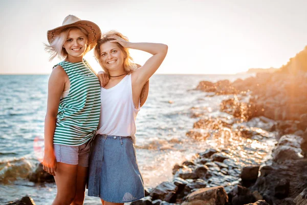 หญิงสาวสองคนที่มีความสุขในหมวกฮิปสเตอร์บนหินบนชายฝั่งทะเล ภูมิทัศน์ฤดูร้อนที่มีผู้หญิง, ทะเล, เกาะและแสงแดดสีส้ม . — ภาพถ่ายสต็อก