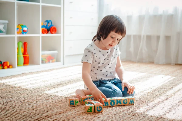 Criança brinca com blocos de madeira com letras no chão na sala uma menina está construindo uma torre em casa ou no jardim de infância . — Fotografia de Stock