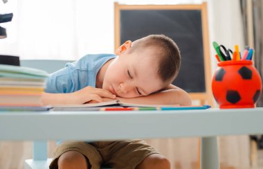 Okullu çocuk sınıfında oturmuş kitap dolu bir masada yatıyor. Uyuyan tembel öğrencileri eğitiyor.