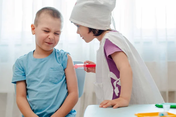 Kinderen spelen in het ziekenhuis. Het meisje is gekleed in een arts 4 jaar oud maakt een injectie aan een jongen 6 jaar oud met een speelgoedspuit in een kamer in een kleuterschool. — Stockfoto