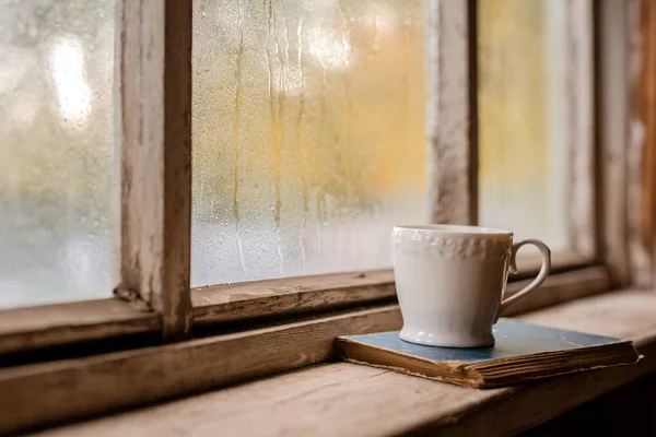 Una taza blanca y libros viejos en el fondo de una ventana húmeda de madera rústica —  Fotos de Stock