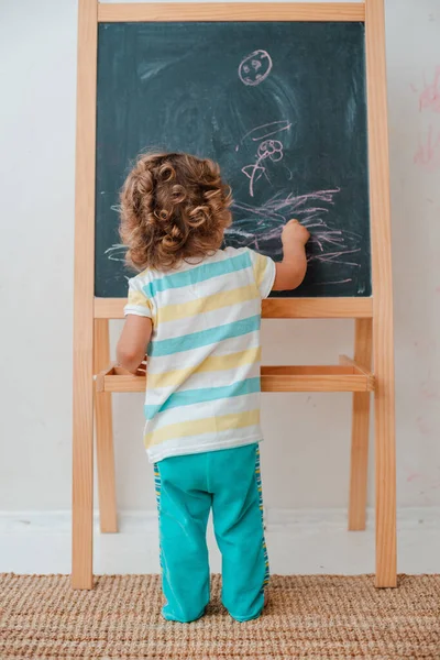 Pequena criança desenha com giz em uma tábua de giz preto em casa no berçário contra uma parede cinza . — Fotografia de Stock