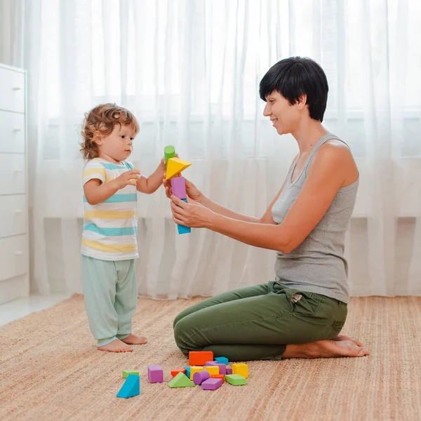 Mother and child play on the floor in the nursery. Mom and a little boy are building a tower of colored blocks.