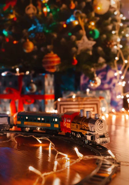 Toy vintage steam locomotive on the floor under a decorated Christmas tree on a background of bokeh lights garland. — Stock Photo, Image