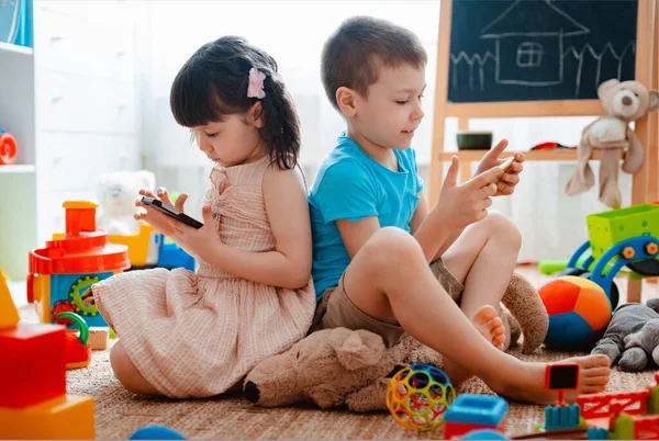 Irmãos crianças irmão e irmã, amigos sentam-se no chão da casa na sala de jogos das crianças com smartphones, destacados dos brinquedos espalhados . — Fotografia de Stock