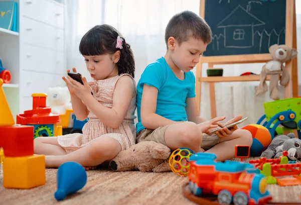 Hermanos niños hermano y hermana, amigos se sientan en el suelo de la casa en la sala de juegos para niños con teléfonos inteligentes, separados de los juguetes dispersos . — Foto de Stock