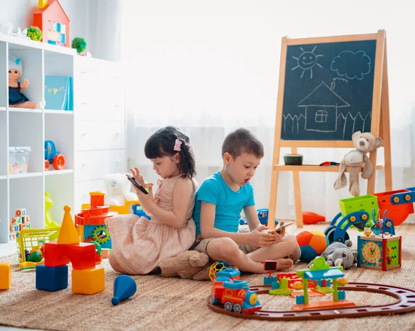 Siblings children brother and sister, friends sit on the floor of the house in the childrens play room with smartphones, detached from the scattered toys. — Stock Photo, Image
