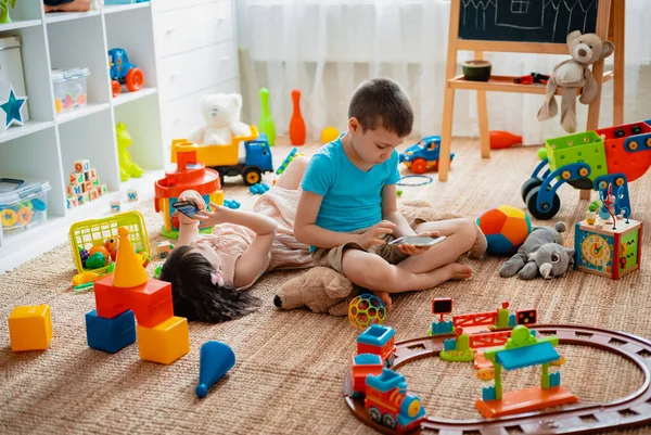 Irmãos crianças irmão e irmã, amigos sentam-se no chão da casa na sala de jogos das crianças com smartphones, destacados dos brinquedos espalhados . — Fotografia de Stock