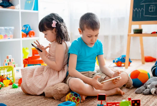 Сидячі діти брат і сестра, друзі сидять на підлозі будинку в дитячій ігровій кімнаті зі смартфонами, відокремленими від розкиданих іграшок . — стокове фото