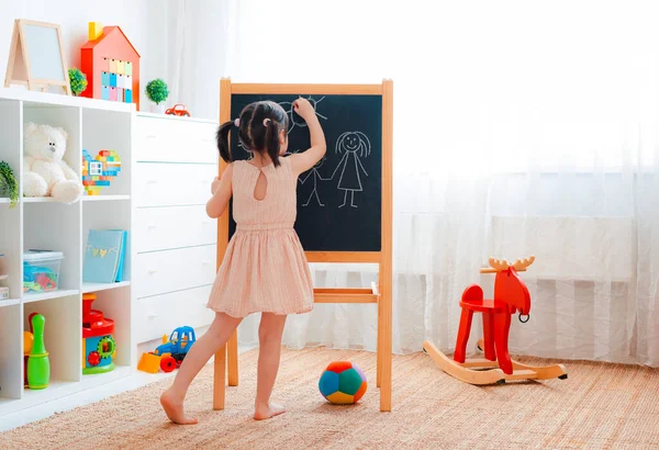 Bir kız elinde tahtayla çocuk odasında duruyor ve tebeşirle bir aile çiziyor.. — Stok fotoğraf