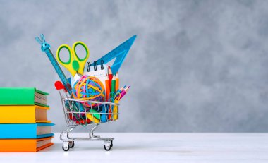 Alış veriş sepetinde renkli okul malzemeleri var. Arka planda da metnin bir kopyası var. Renkli kapaklı bir yığın kitap. Yeni öğretim yılı için okula dönüş kavramı.