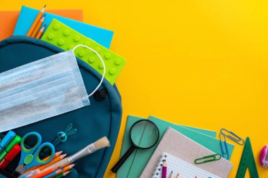 Okul malzemeleri, mavi sırt çantası tıbbi maske sarı arka plan. Okula geri dönmek, pandemik koronavirüs konsepti, düz kat. Yatay. Seçici odaklanma. Yukarıdan bak. Boşluğu kopyala.