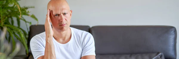 Desperate young man keeping his hand on forehead while sitting on the couch at home — Stock Photo, Image