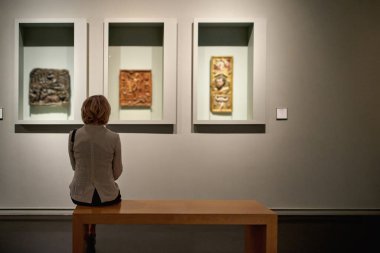 Renkli tabloların önünde bir sanat galerisinde oturan kadının dikiz görüntüsü