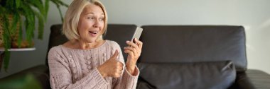Kanepede otururken cep telefonu kullanan yaşlı bir kadın. Yaşlı kadın koltukta oturuyor ve bir telefon mesajı yolluyor. Akıllı telefon kullanmayı öğrenen güzel yaşlı bir kadının portresi..