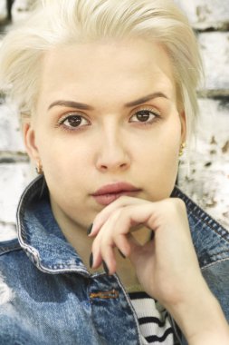 Genç güzel sarışın kız Kot Mavi giysili yaz öğleden sonra tuğla duvarı ayakta.