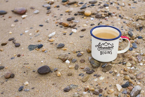 A metal enameled travel mug with pure natural drinking Baikal water on the rocks and sand of the shore of Lake Baikal in the summer in a sunny clear day.