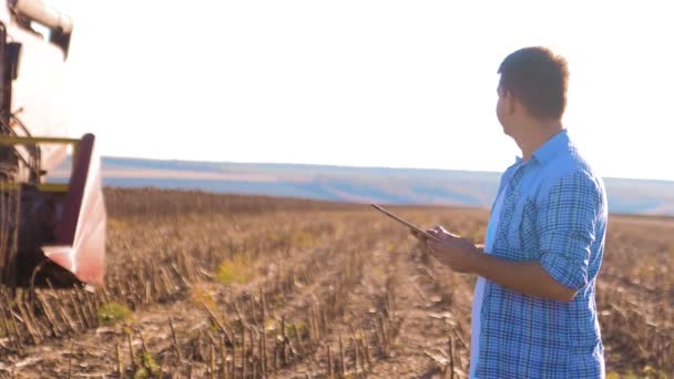 Фермер работает умный фермер читать или анализировать урожай подсолнечника отчет в планшетном компьютере на сельскохозяйственном поле с винтажным тоном на солнечном свете. комбайн вспахивает поле культуры — стоковое видео