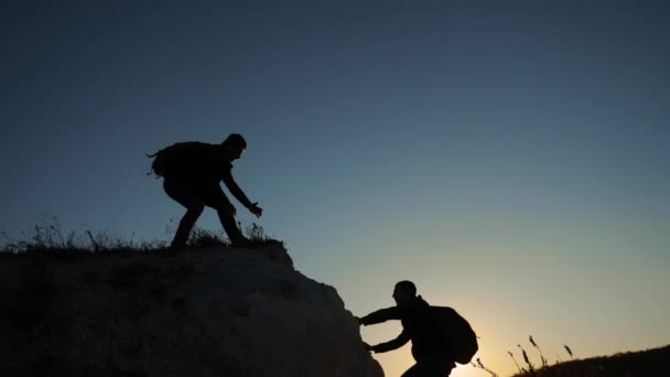 Silhouette klettern zwei Männer in Teamarbeit einen Berg hinauf. Wandertouristen wandern Abenteuerkletterer bei Sonnenuntergang den Berg hinauf. Zeitlupenvideo. Wanderer gewinnen Sieg auf der Schanze — Stockvideo