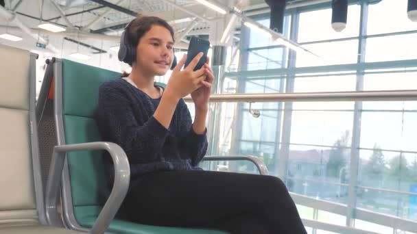 airport waiting hall room for a flight by plane. young happy teen girl in headphones listening to the music on smartphone chatting communicates in the messenger. teenager girl in social networks