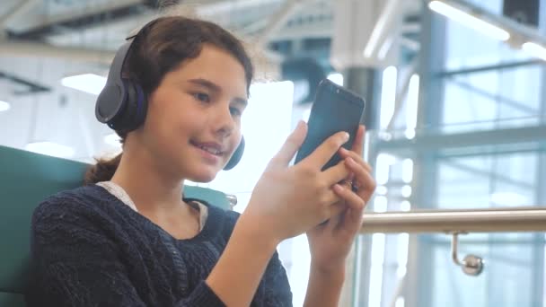 airport waiting hall room for a flight by plane. young happy teen girl in headphones listening to lifestyle the music on smartphone chatting communicates in the messenger. teenager girl in social